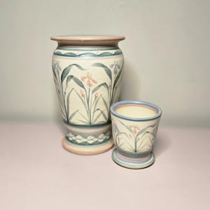 May include: Two ceramic vases, one tall and one small, with a floral iris design. The vases are cream-colored with blue, green, and peach accents. The tall vase has a wide base and a flared rim. The smaller vase has a pedestal base.