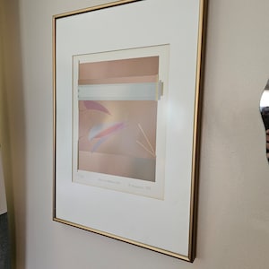 May include: A framed print of an abstract art piece with a gold frame. The print features a layered design of pink, white, and blue colours. The artist's name and date are printed in the bottom right corner of the print.
