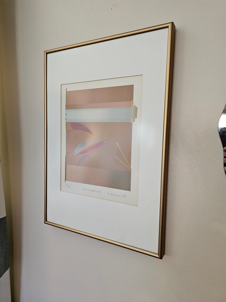 May include: A framed print of an abstract art piece with a gold frame. The print features a layered design of pink, white, and blue colours. The artist's name and date are printed in the bottom right corner of the print.