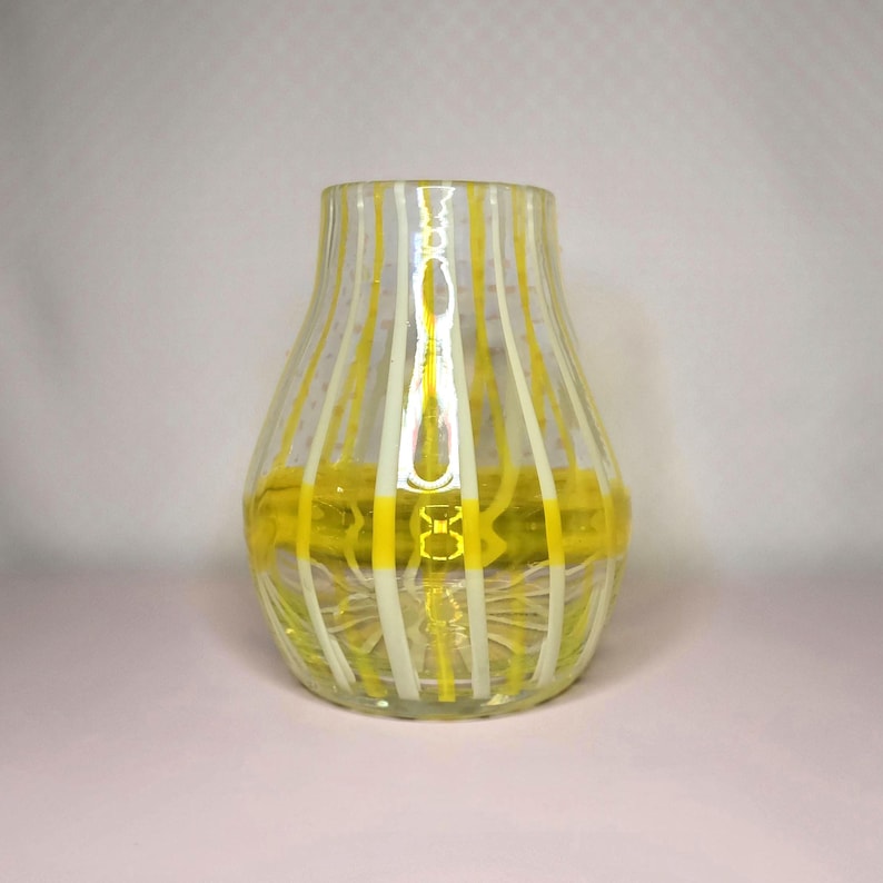 May include: A clear glass vase with vertical yellow and white stripes. The vase has a rounded shape with a wide base and a narrower neck. The interior of the vase is filled with a yellow substance, adding a pop of color.