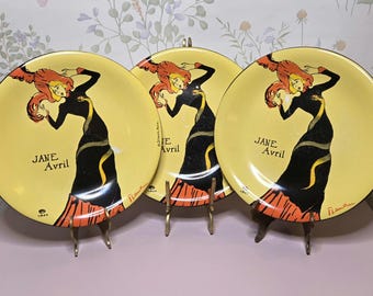 May include: Three round ceramic plates featuring a yellow background and a black and orange illustration of a woman in a long dress. The text Jane Avril is printed on each plate, along with the signature Lautrec and the year 1893 in black ink.