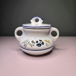 May include: White ceramic sugar bowl with a lid and two handles. The bowl is decorated with blue floral patterns and blue trim. The lid has a small handle with a hole. The bowl is sitting on a pink surface.