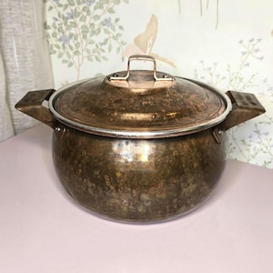 May include: A round, hammered copper pot with a matching lid and two side handles. The metal surface displays a weathered, dark brown patina with visible texture and metallic highlights. The lid features a central handle for easy removal.