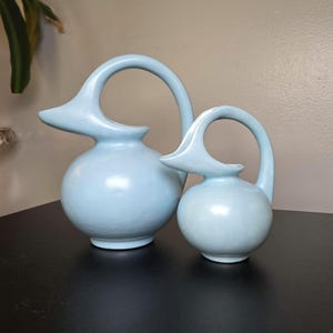 May include: Two light blue ceramic pitchers with rounded bodies and elongated, curved handles that extend from the top of the spout to the base. The pitchers feature a smooth, matte finish and a distinct, bird-like silhouette with a pointed spout.