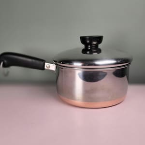 May include: A small stainless steel saucepan with a lid and a black handle. The bottom of the pan has a copper band. The lid has a black knob. The pan is sitting on a pink surface.