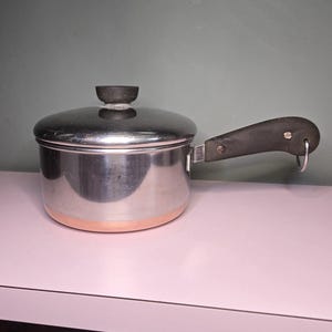 May include: A stainless steel saucepan with a lid and a black handle. The pot has a copper-colored bottom and a small, round black handle on the lid. The handle has a metal ring for hanging. The pot is sitting on a pink surface.