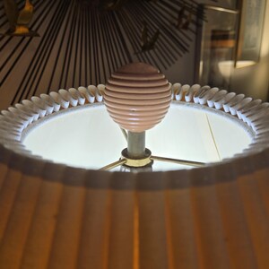 May include: A close-up of a lamp with a pleated, tan lampshade. The lamp has a light pink, ribbed sphere finial. The lamp's base is gold-coloured, and the shade is trimmed with a white, scalloped edge. The lamp is lit from within.