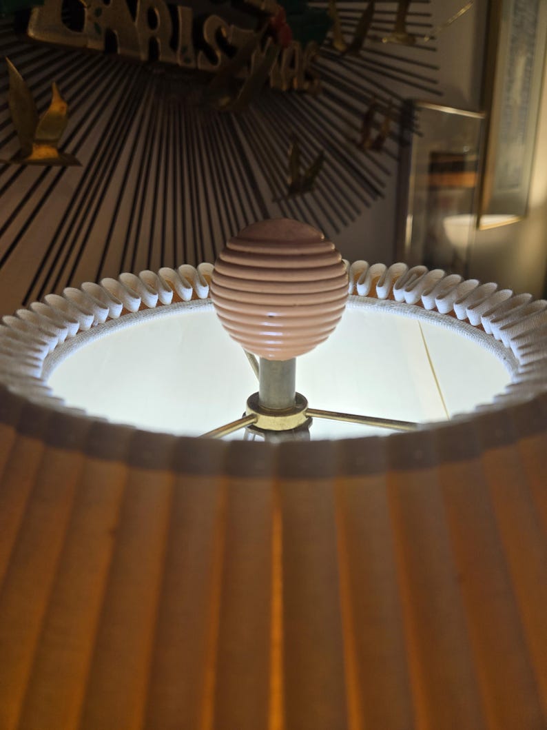 May include: A close-up of a lamp with a pleated, tan lampshade. The lamp has a light pink, ribbed sphere finial. The lamp's base is gold-coloured, and the shade is trimmed with a white, scalloped edge. The lamp is lit from within.
