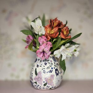 May include: A floral arrangement in a decorative vase. The vase is white with a blue and purple floral pattern. The flowers are a mix of white, pink, and orange blooms with green foliage. The background is a soft, patterned design.