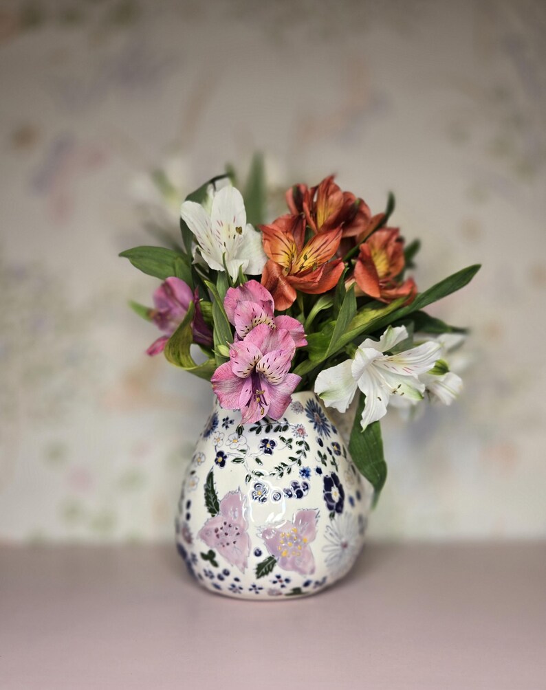 May include: A floral arrangement in a decorative vase. The vase is white with a blue and purple floral pattern. The flowers are a mix of white, pink, and orange blooms with green foliage. The background is a soft, patterned design.