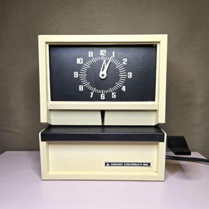 May include: Cream-colored vintage time clock with a black clock face and white numerals. The clock face shows the time as 1:00. The clock has a rectangular shape with a drawer and the text "AMANO CINCINNATI INC."