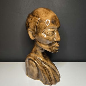 May include: A brown ceramic bust of a person with closed eyes, a prominent nose, and full lips. The sculpture has a smooth, glossy finish and is set against a dark gray background.