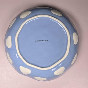 May include: A light blue ceramic plate with a wavy edge and white cloud-shaped accents. The plate has a light blue interior and a white rim. The text "by ANTHROPOLOGIE" is printed on the interior.