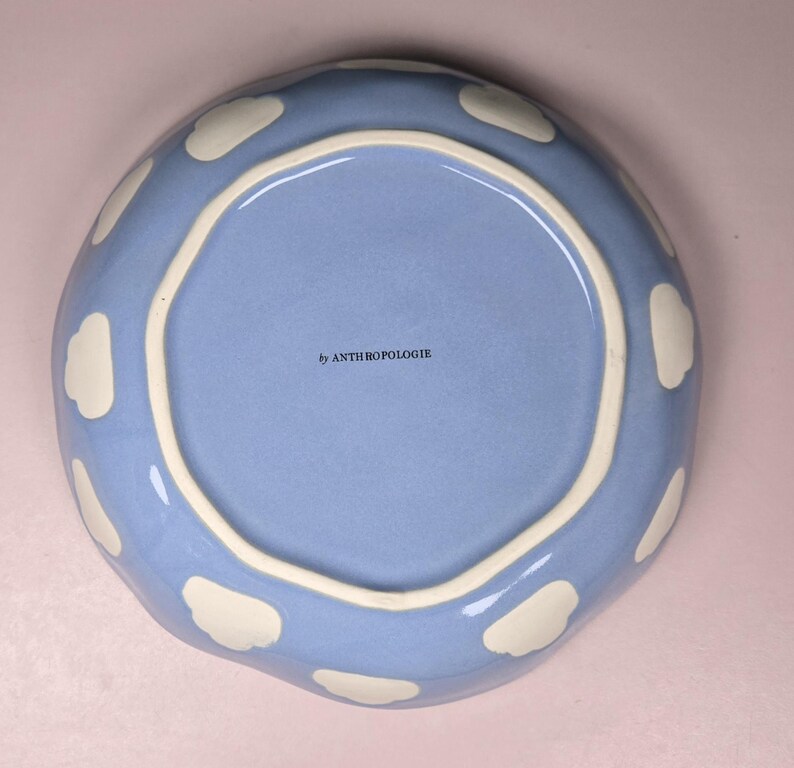 May include: A light blue ceramic plate with a wavy edge and white cloud-shaped accents. The plate has a light blue interior and a white rim. The text "by ANTHROPOLOGIE" is printed on the interior.
