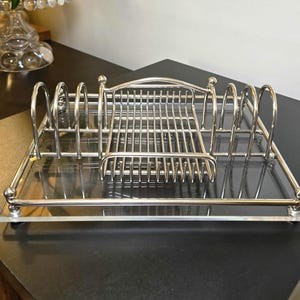 May include: A silver-toned metal toast rack featuring a rectangular base with a central grid compartment and four arched dividers on each side. The structure is mounted on a clear, rectangular acrylic tray with small feet at each corner.