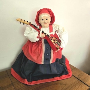 May include: A folk art doll dressed in traditional Russian attire, holding a stringed instrument. The doll wears a red headscarf, a white blouse, and a red and blue dress. The instrument is red and gold with a wooden neck.