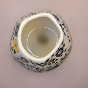 May include: A white ceramic vase with a unique, irregular shape. The vase is adorned with a pattern of yellow, blue, and purple floral designs. The interior is a light beige color, and the vase sits on a light pink surface.