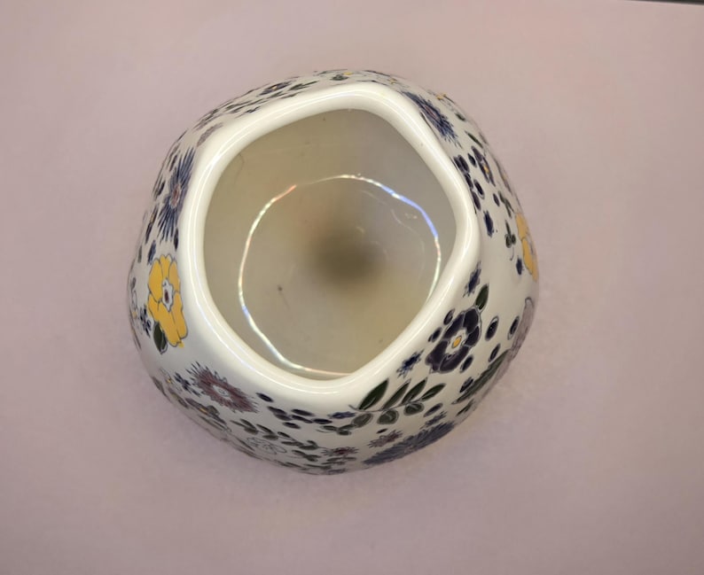 May include: A white ceramic vase with a unique, irregular shape. The vase is adorned with a pattern of yellow, blue, and purple floral designs. The interior is a light beige color, and the vase sits on a light pink surface.