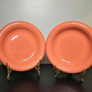 May include: Two round, solid coral-colored ceramic plates with wide rims. Each plate is displayed upright on a small, gold-toned metal wire stand. The plates have a smooth, glossy finish and a simple, circular design with no patterns or decorations.