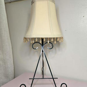 May include: A vintage table lamp with a cream-colored lampshade and a black metal base. The lampshade has a scalloped edge with dangling crystal beads. The base is an A-frame design with decorative scrollwork.