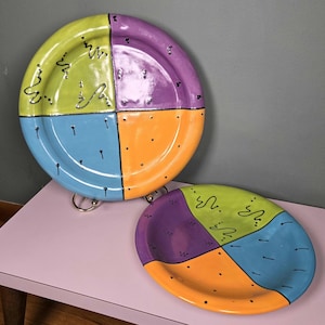 May include: Two colorful ceramic plates. The round plate is divided into four sections: green, purple, blue, and orange. The oval plate has the same color scheme. Both plates have black squiggles and dots.