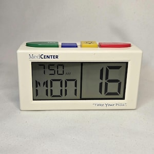 May include: A white MedCenter digital clock with a green, blue, yellow, and red button. The clock displays the time as 7:50 AM on Monday and the number 16. The text "Take Your Pills" is below the number 16.