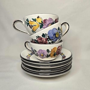 May include: A stack of white ceramic teacups and saucers. Each cup and saucer is decorated with colorful floral designs in yellow, purple, and red. The rims and handles are outlined in black.