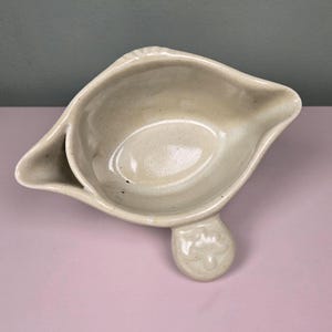 May include: A cream-colored ceramic gravy boat with a smooth, glossy finish. The vessel features a wide, oval-shaped basin, a pointed pouring spout on one side, and a small, rounded handle extension with a subtle embossed fleur-de-lis design.