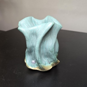 May include: A small ceramic vase with a textured, wavy surface in shades of teal and blue-green. The base features a subtle tan or beige coloration, and a single small, round, light-colored bead is embedded near the bottom of the exterior wall.