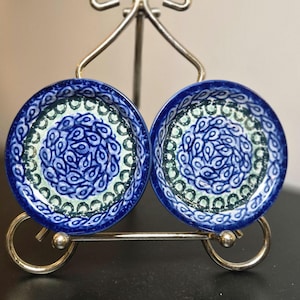 May include: Two small, round ceramic dishes featuring a dark blue rim and a central pattern of blue teardrop shapes on a light green background. The dishes are displayed side-by-side on a silver-toned metal wire stand with a curved base and top.