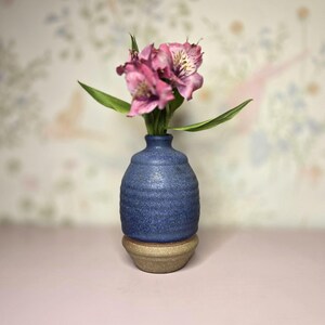 May include: A small, blue ceramic vase with a textured surface, holding pink flowers with green leaves. The vase sits atop a smaller, tan-colored base, creating a layered effect. The background features a soft, floral pattern.