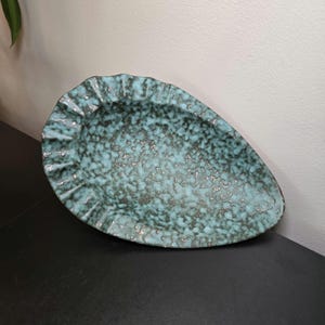 May include: A teardrop-shaped ceramic dish featuring a textured, mottled glaze in shades of turquoise and dark charcoal gray. The rim of the dish has a distinct, ruffled, or pleated edge, creating a decorative border around the central basin.