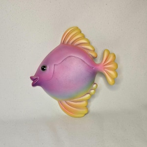 May include: A decorative fish figurine in shades of pink, yellow, and blue. The fish has a rounded body, a small mouth, and large fins. The fins and tail are yellow, while the body transitions from pink to blue.