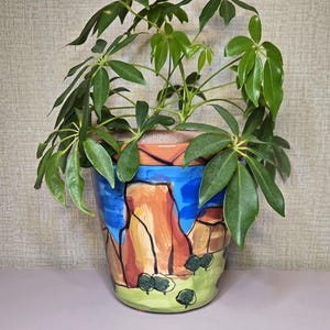 May include: A potted plant with vibrant green leaves in a hand-painted ceramic pot. The pot features a landscape design with red rock formations, blue sky, and green foliage. The pot is approximately 8 inches tall.