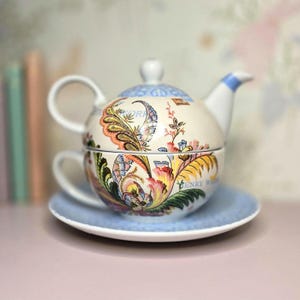 May include: A decorative tea set featuring a teapot and teacup stacked together on a light blue saucer. The set is adorned with a colorful floral pattern in shades of orange, yellow, and blue. The teapot has a white handle and spout.