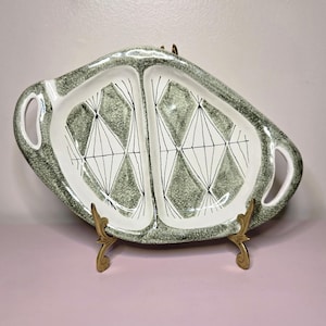 May include: A vintage divided serving platter with a white interior and a gray speckled exterior. The platter features a geometric diamond pattern in black and white and is supported by two decorative gold-colored stands. It has two handles.