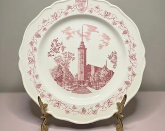 May include: A white decorative plate with a scalloped edge and a red illustration of a building and trees. The plate has a red vine and leaf border and is displayed on a gold-colored stand.