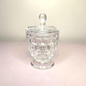 May include: A clear, faceted glass jar with a lid. The jar has a pedestal base and a diamond pattern on the body. The lid has a small, rounded handle. The jar is likely used for storing small items.