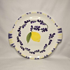 May include: A white ceramic serving platter with a hand-painted lemon and lavender design. The yellow lemon has a green leaf, and the lavender is purple. The rim has yellow and blue stripes and two handles.