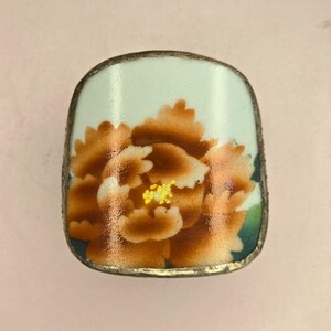 May include: A curved, rectangular metal ring component featuring a glossy enamel surface. The design displays a large, detailed brown peony flower with yellow stamen accents against a pale mint green background, framed by a thin silver-toned metal border.