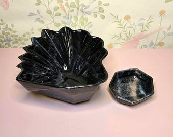 May include: A black ceramic bowl with a pleated, fan-like design and a matching small hexagonal dish. Both pieces feature a glossy finish with subtle white marbling or highlights throughout the dark surface, creating a cohesive set of vessels.