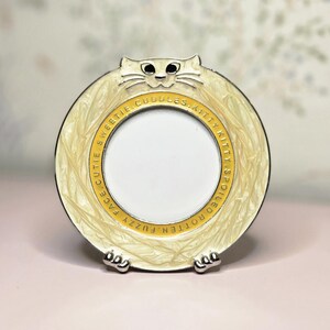 May include: A cream-colored cat-shaped picture frame with a round opening. The frame has a textured border and a yellow inner ring with text. The cat has black eyes and silver paws.