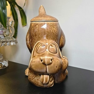 May include: A brown ceramic cookie jar shaped like a dog with long ears, a protruding tongue, and a textured coat. The dog is in a seated position with its head tilted forward, and the top portion of the jar forms a removable lid with a pointed top.