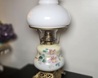 May include: Antique white glass lamp with a ruffled top and floral painted base. The lamp features a brass-colored metal frame and base. The base is decorated with pink and green flowers. A classic decorative lighting piece.