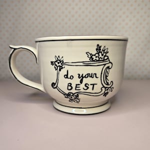 May include: Cream-colored ceramic mug with a black handle and rim. The mug features the phrase "do your BEST" in black script within a decorative frame, accented with floral motifs. The mug sits on a light pink surface.