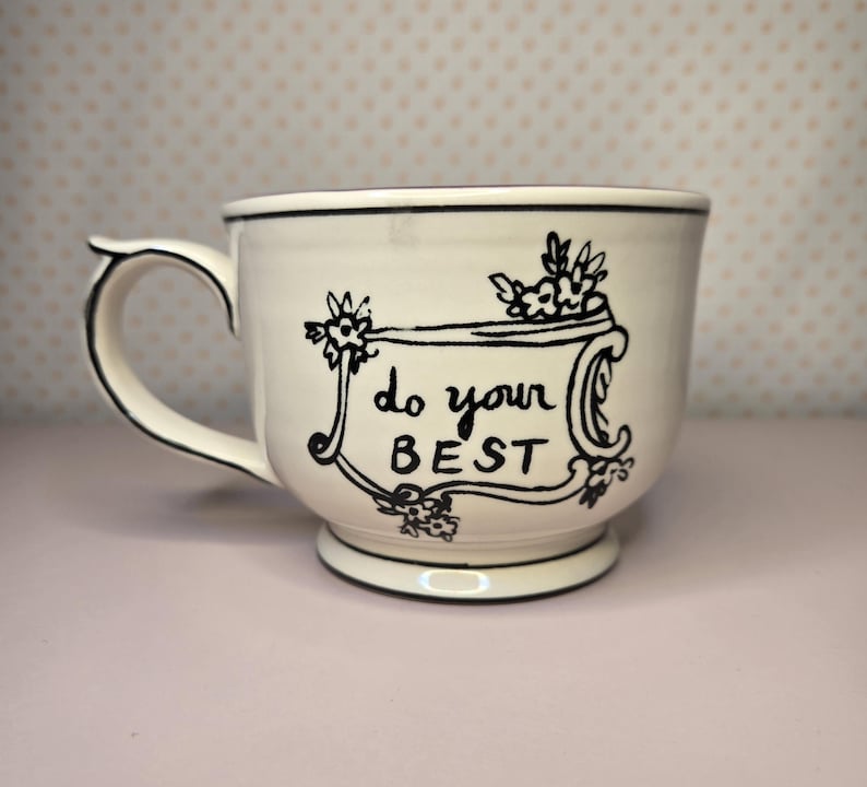 May include: Cream-colored ceramic mug with a black handle and rim. The mug features the phrase "do your BEST" in black script within a decorative frame, accented with floral motifs. The mug sits on a light pink surface.