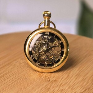 May include: A gold-toned pocket watch featuring a skeleton dial that reveals the intricate internal mechanical movement. The watch face has a black outer ring with white Roman numerals and thin black hands, topped with a gold winding crown.