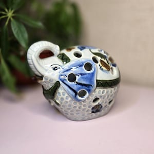 May include: A ceramic elephant figurine with a blue and white patterned design. The elephant has a raised trunk and a blue decorative element with several holes. The figurine is decorated with green and brown accents.