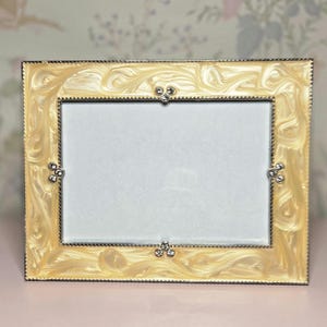 May include: Rectangular photo frame with a cream-colored, swirled design. The frame has a silver-colored trim and decorative accents at each corner. The frame is empty, ready to display a photograph. A decorative item for home decor.