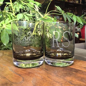 May include: Two dark-tinted whiskey glasses with etched designs. One glass features a motorcycle, the other displays the letters "RS". The glasses have a clear base and sit on a wooden surface, with green foliage in the background.